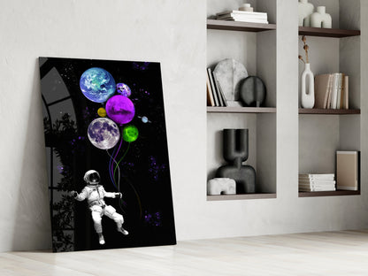 Astronaut holding colorful planet balloons against black starry background glass wall art leaning against white wall beside shelving.