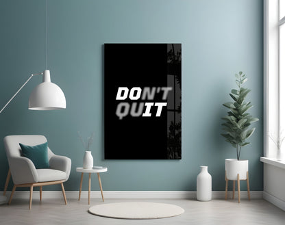 Black glass wall art displaying "DON'T QUIT" text in white, mounted in modern room with teal walls, white chair, and potted plants.