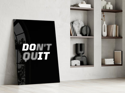 Black glass wall art with white "DON'T QUIT" text leaning against white wall beside wooden shelving with decorative objects.