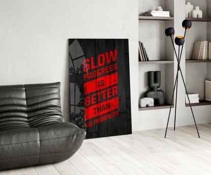 Modern living room with black leather chair, leaning glass wall art featuring red text "Slow Progress Is Better Than No Progress" on dark background, black tripod lamp, and shelving unit with books and decor.