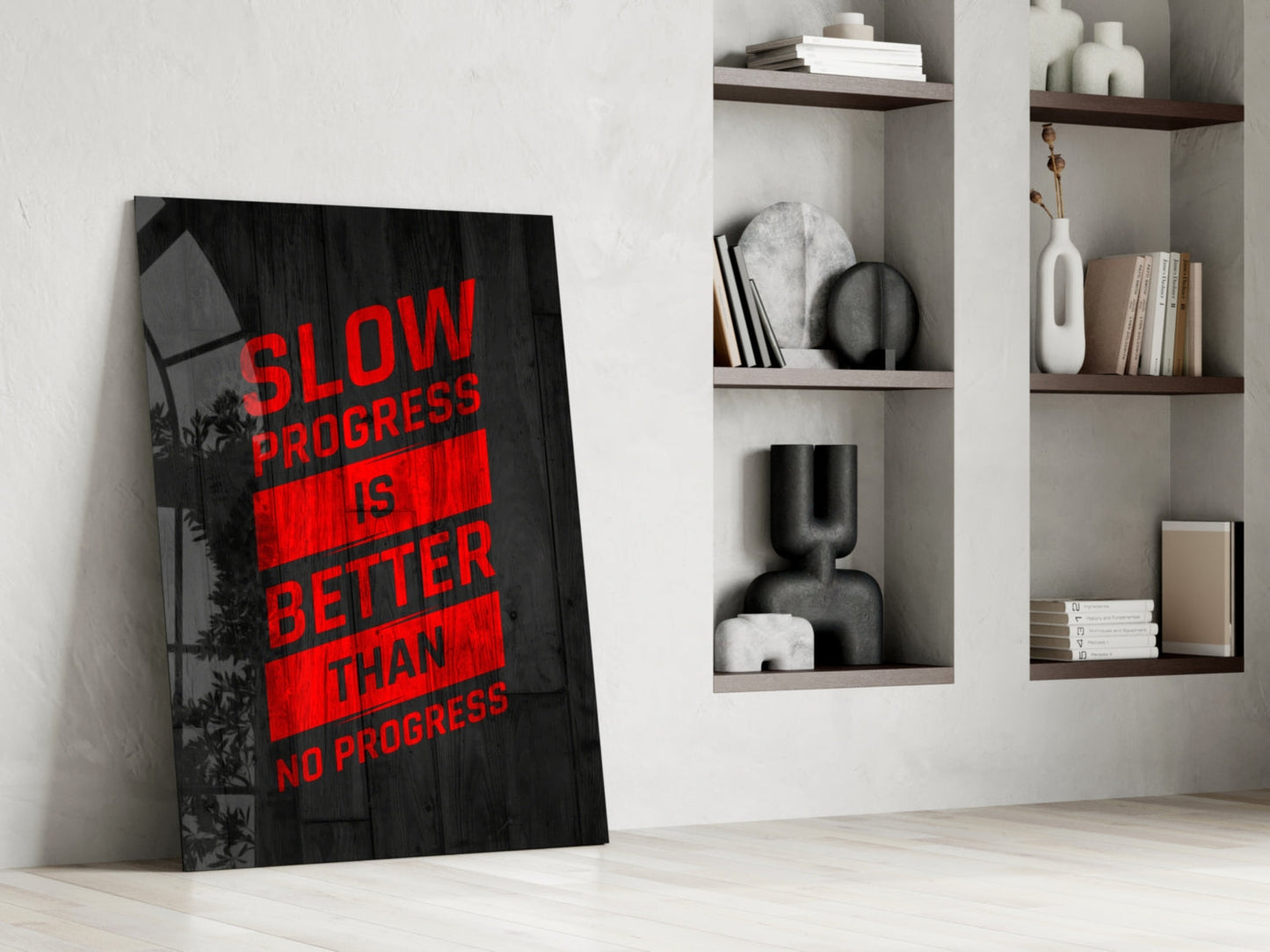 Black glass wall art with red text reading "Slow Progress Is Better Than No Progress" leaning against white wall beside wooden shelves.
