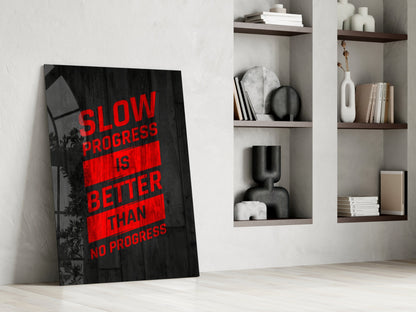 Black glass wall art with red text reading "Slow Progress Is Better Than No Progress" leaning against white wall beside wooden shelves.