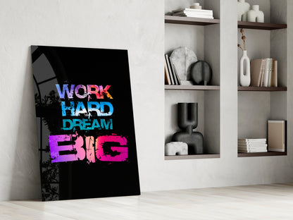 Colorful glass wall art leaning against white wall displaying "WORK HARD DREAM BIG" text in purple, blue, and pink on black background, with white shelving unit beside it.