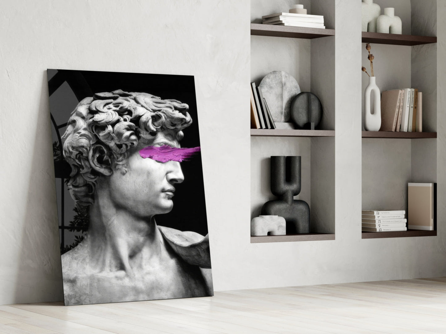 Modern interior room with leaning glass wall art featuring classical bust sculpture with purple accent across eyes, beside white shelving unit holding books and decorative objects.