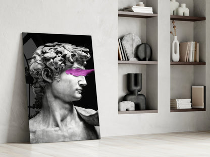 Modern interior room with leaning glass wall art featuring classical bust sculpture with purple accent across eyes, beside white shelving unit holding books and decorative objects.