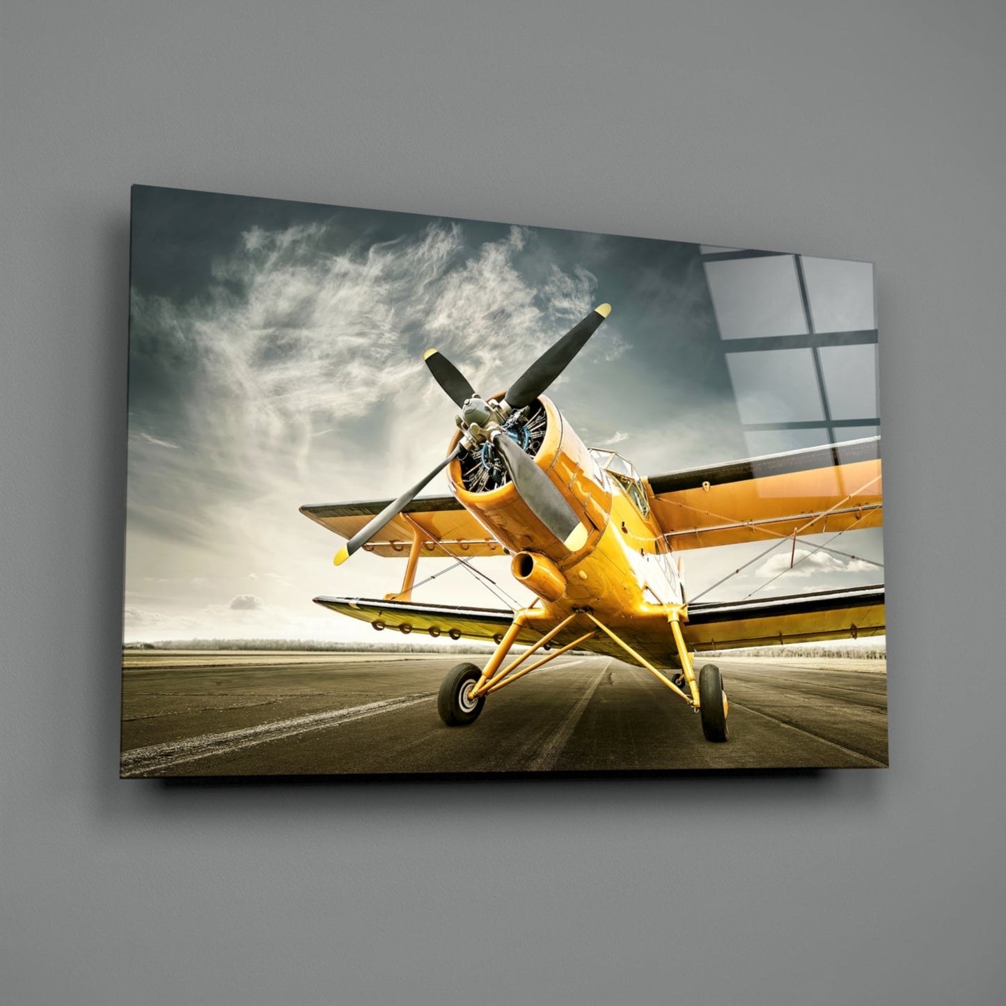 Yellow vintage biplane on runway against dramatic cloudy sky, displayed as framed glass wall art on gray wall.