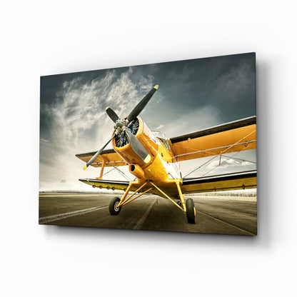 Vintage yellow biplane on runway viewed from front quarter angle against dramatic cloudy sky, displayed on glass wall art panel.