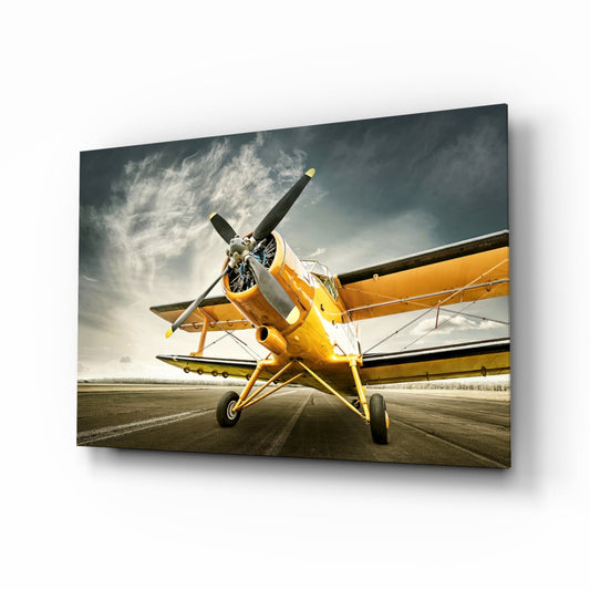 Vintage yellow biplane on runway viewed from front quarter angle against dramatic cloudy sky, displayed on glass wall art panel.