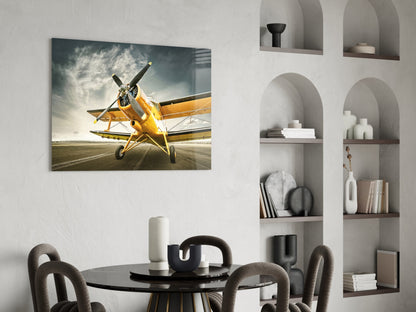 Yellow vintage biplane on runway with dramatic cloudy sky, displayed as glass wall art in modern dining room setting.