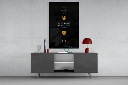 Black glass wall art with gold sun, heart, and arrow symbols above white text reading "By His Wounds We Are Healed - 1 Peter 2:24" displayed on gray media console.