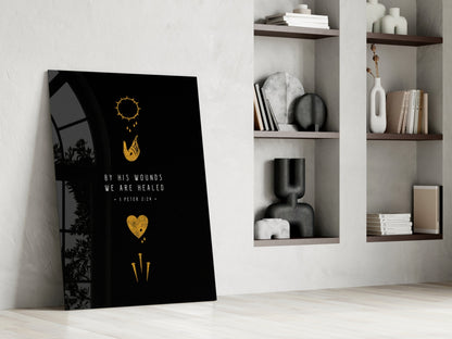 Black glass wall art leaning against white wall displaying gold sun, hand, heart, and sword symbols with "By His Wounds We Are Healed" text, styled home setting with built-in shelving.