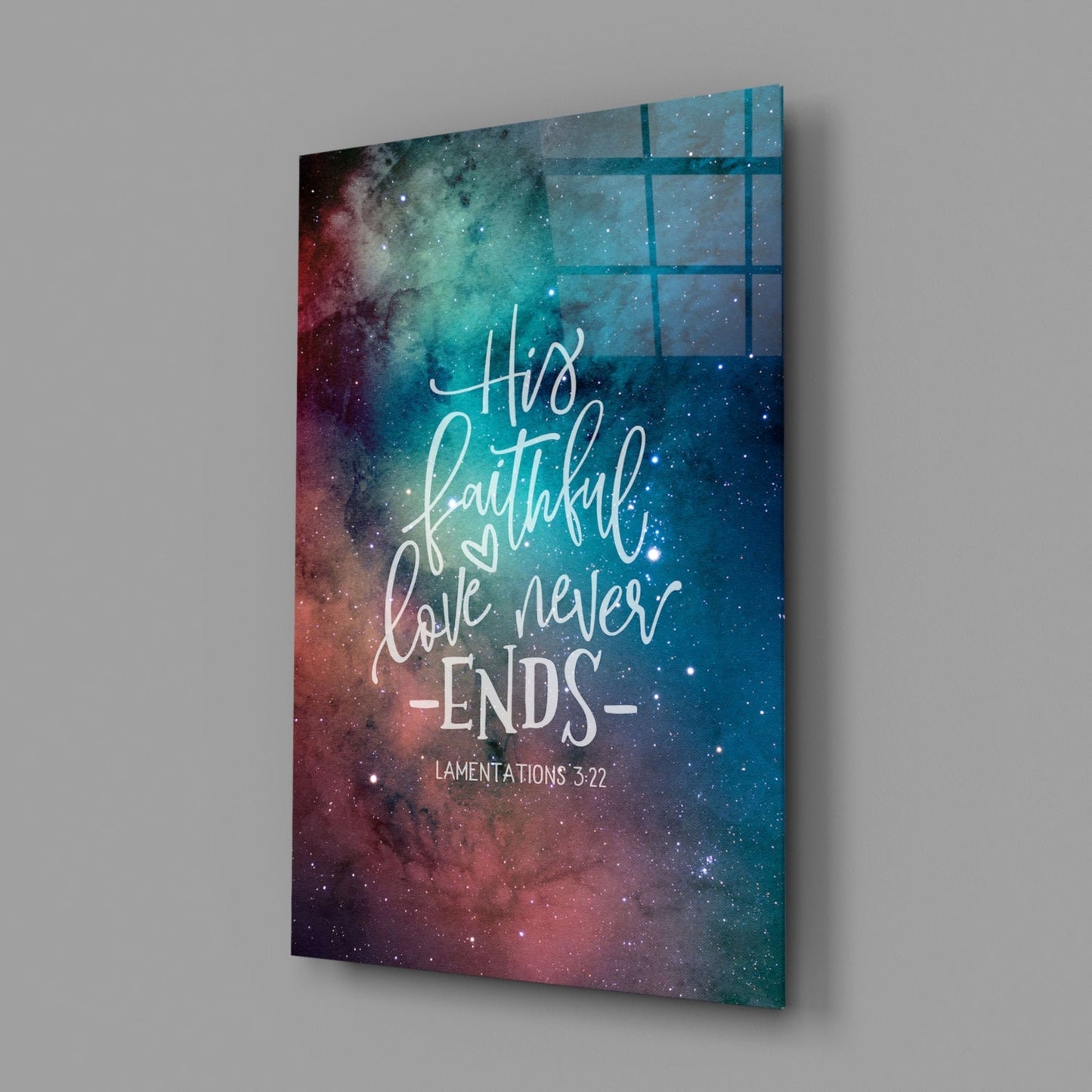Glass wall art panel displaying galaxy background with red and teal nebula colors, featuring white script text reading "His faithful love never ends" and "Lamentations 3:22" citation, shown at an angle against gray wall.