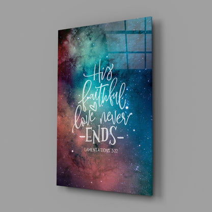 Glass wall art panel displaying galaxy background with red and teal nebula colors, featuring white script text reading "His faithful love never ends" and "Lamentations 3:22" citation, shown at an angle against gray wall.