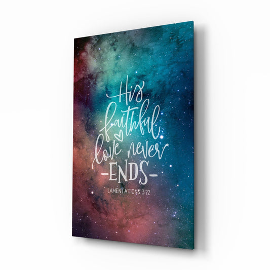 Angled perspective view of glass wall art with galaxy background in teal and burgundy tones displaying white script text reading "His faithful love never -ENDS-" and "LAMENTATIONS 3:22".