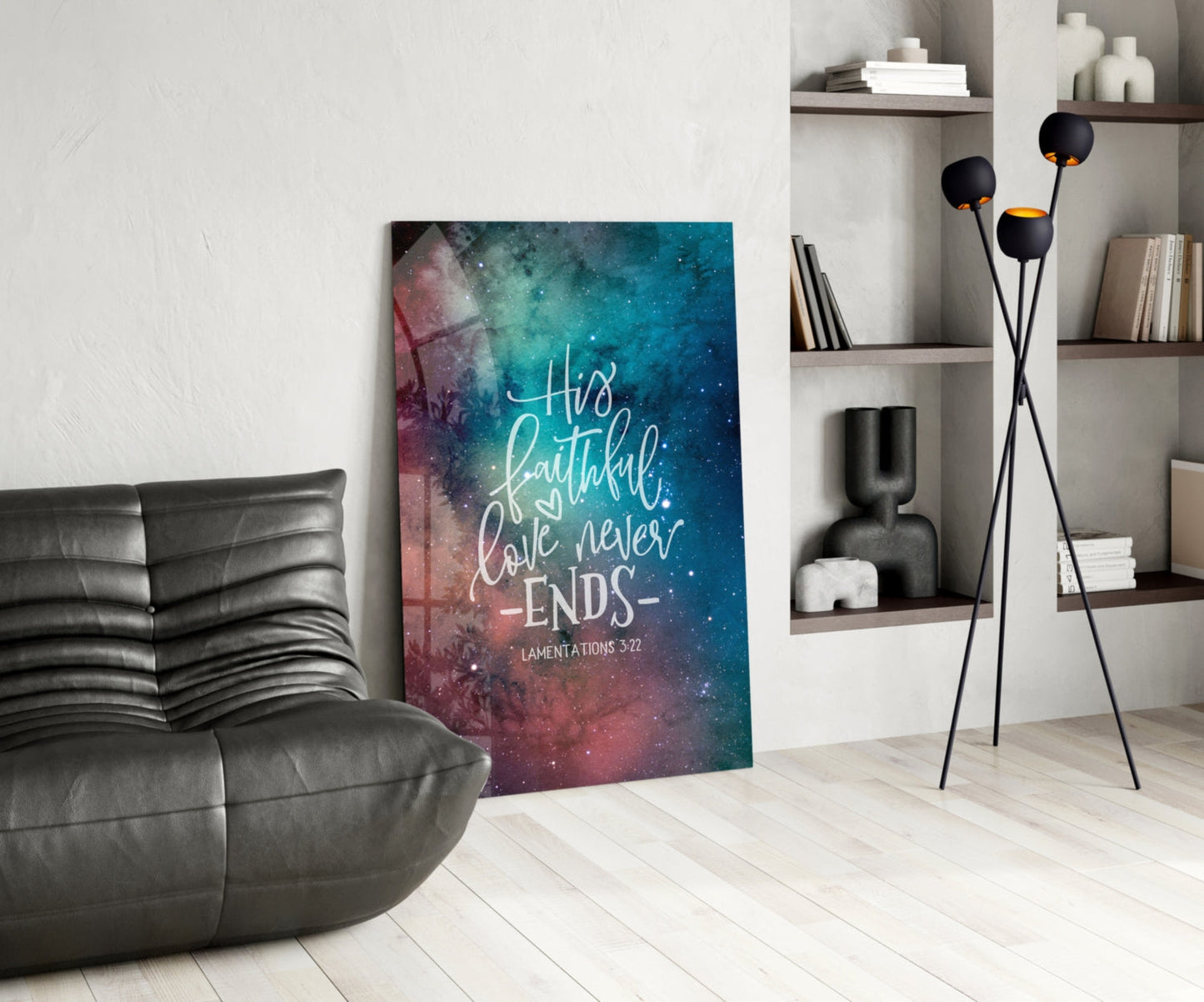 Modern living room interior featuring a teal and pink galaxy-printed glass wall art with white script reading "His faithful love never ends" and "Lamentations 3:22" positioned against a white wall beside a black leather chair.