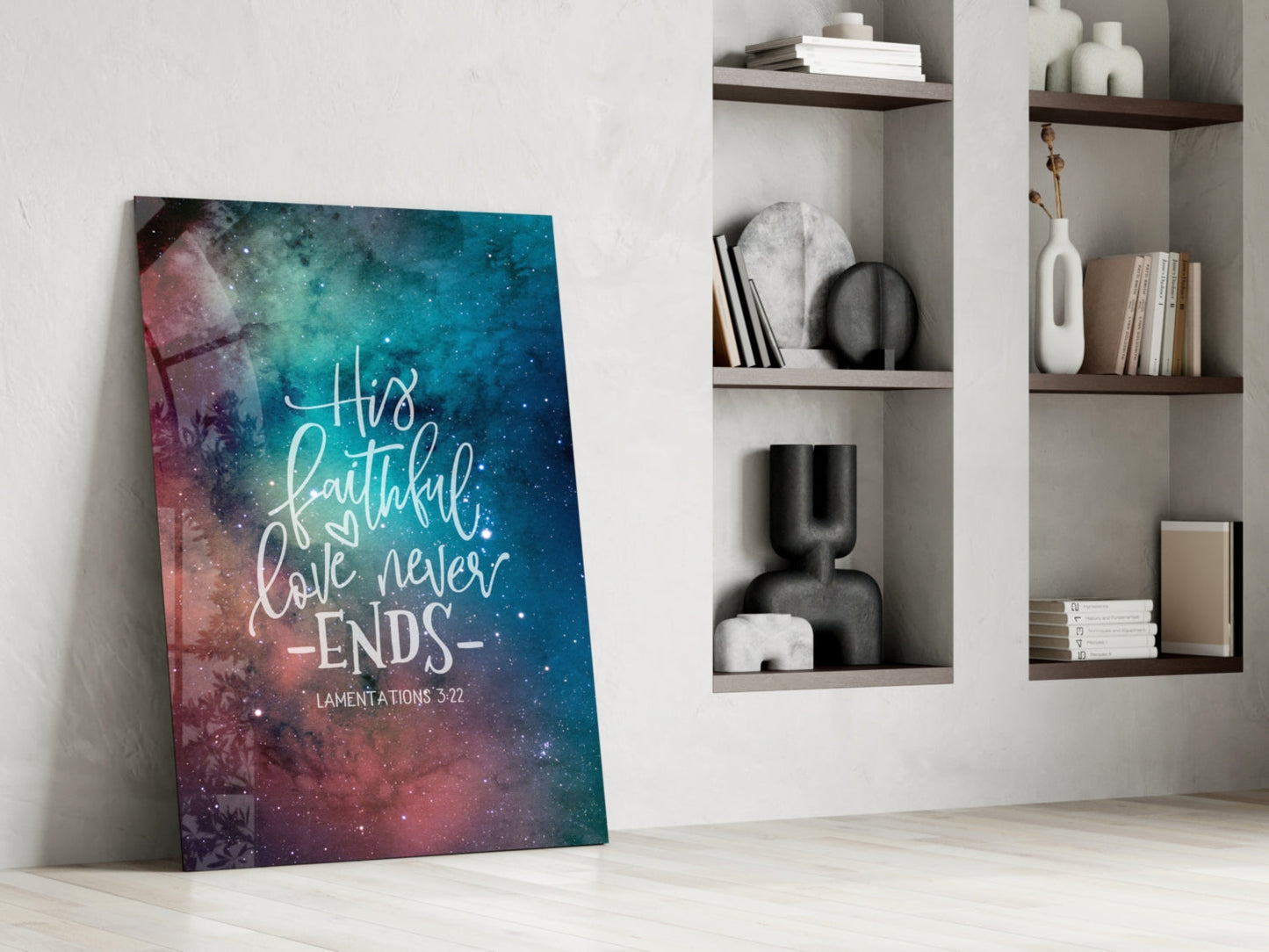 Glass wall art with galaxy background in teal and pink displaying white script text "His faithful love never ends" and "Lamentations 3:22" leaning against white wall with shelving.
