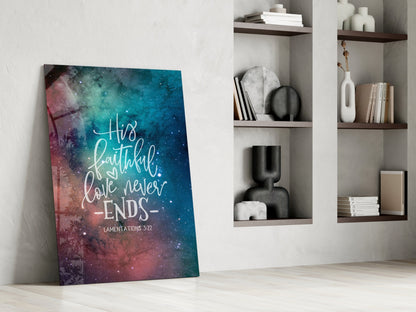 Glass wall art with galaxy background in teal and pink displaying white script text "His faithful love never ends" and "Lamentations 3:22" leaning against white wall with shelving.
