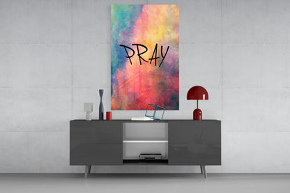 Colorful watercolor glass wall art displaying "PRAY" text in black handwriting, displayed above a dark gray media console in a modern interior setting.