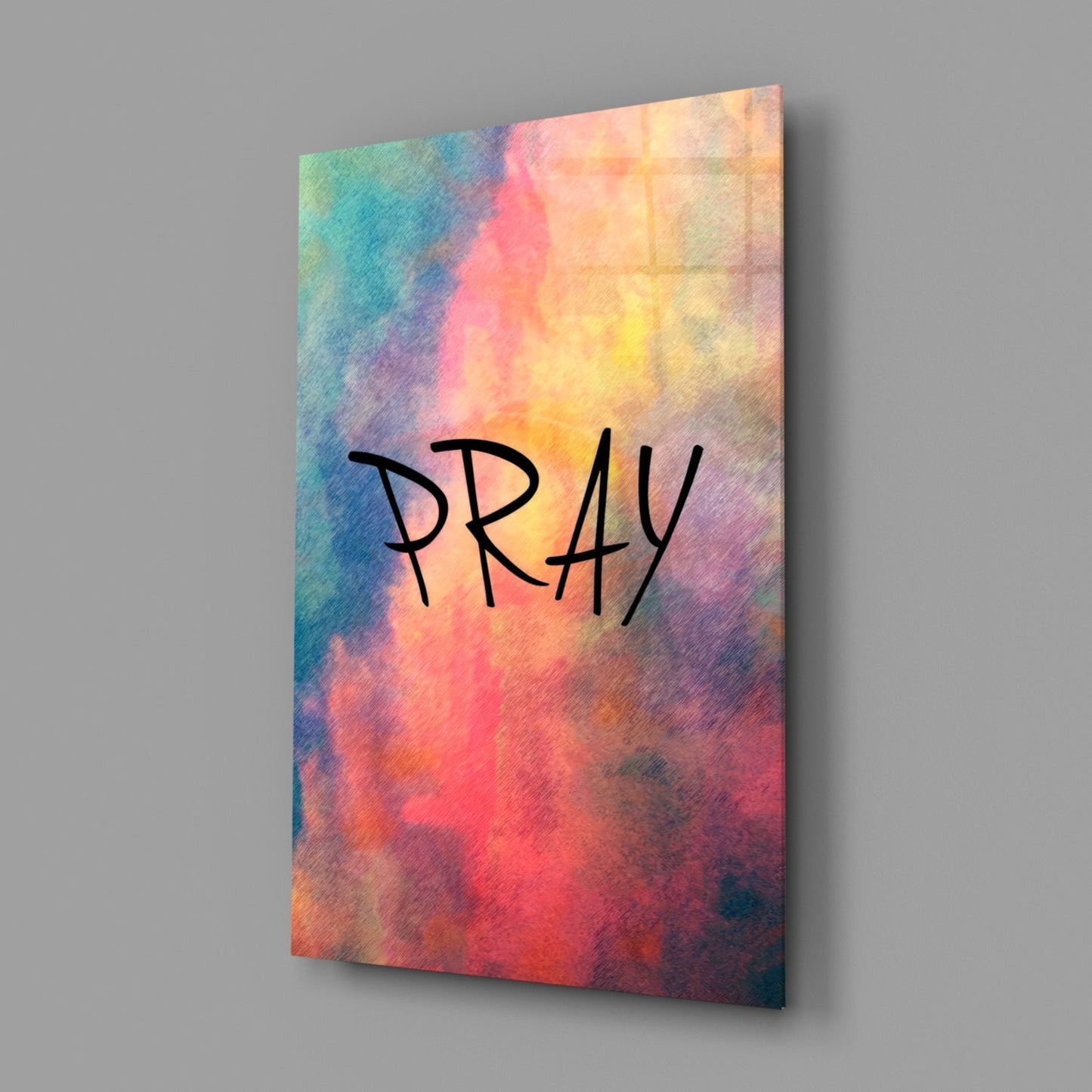 Glass wall art panel with watercolor gradient background in teal, pink, yellow, and red tones displaying "PRAY" in black handwritten text, angled three-quarter view.