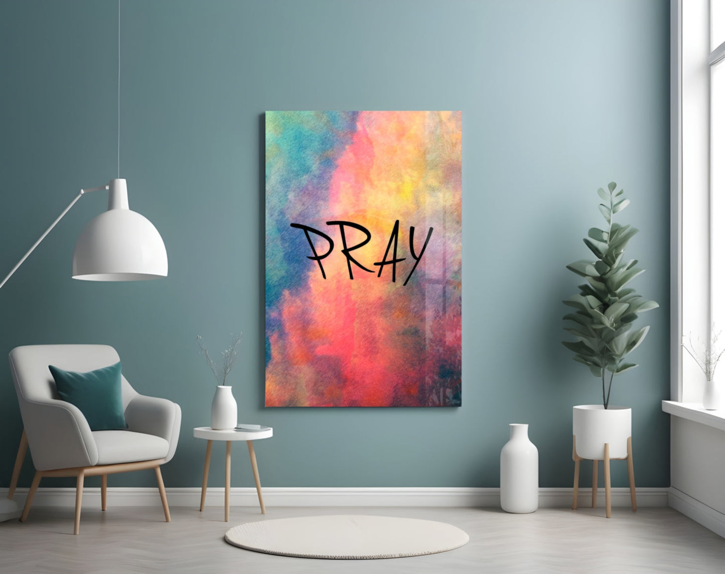 Colorful watercolor glass wall art displaying "PRAY" text in black handwriting, mounted on teal wall in modern living room setting with chair and plants.