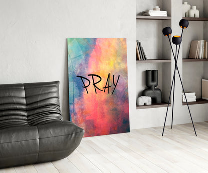 Colorful watercolor glass wall art displaying "PRAY" text in black lettering, displayed in a modern living room setting with leather furniture and shelving.