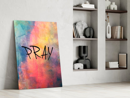 Colorful watercolor gradient glass wall art leaning against white wall displaying "PRAY" text, with shelving and decorative objects visible behind.