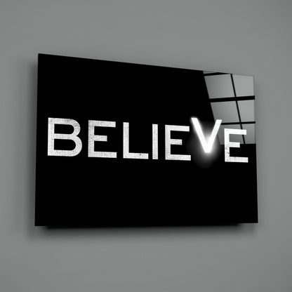 Black glass wall art panel with white "BELIEVE" text and illuminated checkmark, displayed at angle against gray background.