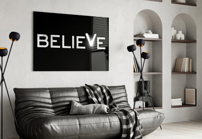 Black glass wall art with white "BELIEVE" text displayed above a gray leather sofa in a modern living room.