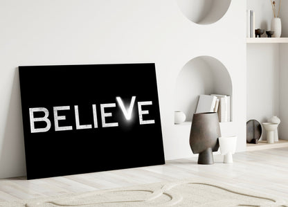 Black glass panel with white "BELIEVE" text leaning against white wall in minimalist room setting.