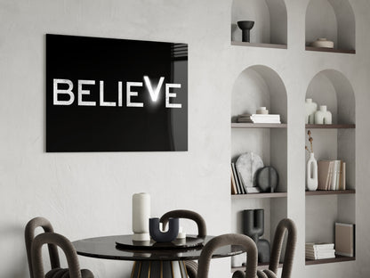 Black glass wall art with white "BELIEVE" text displayed above a round black dining table in a minimalist white interior with built-in shelving.