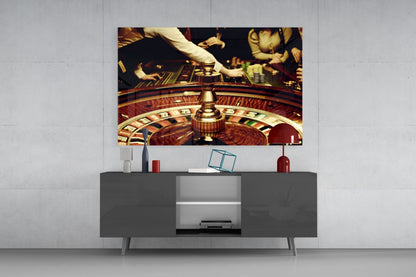 Glass wall art depicting a roulette wheel with casino players and chips mounted above a gray media console in a modern interior setting.