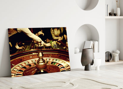 Gamble Glass Wall Art leaning against white wall in minimalist room, showing roulette wheel and casino scene in warm gold and brown tones.