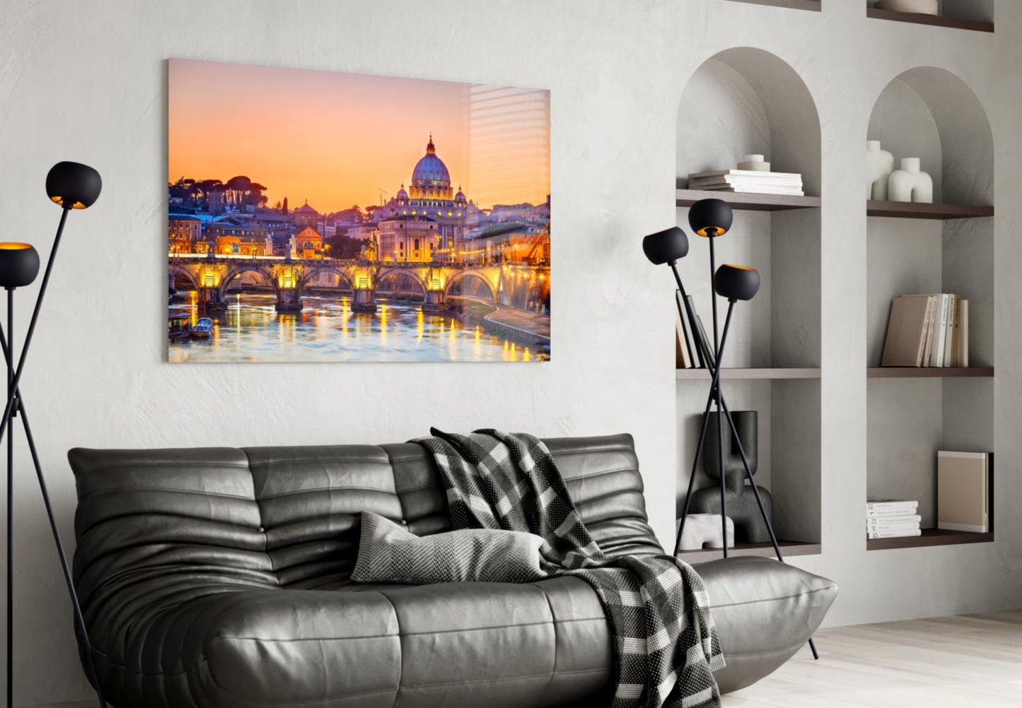 Glass wall art depicting St. Peter's Basilica at sunset with illuminated bridge and water reflection, displayed above black leather sofa in modern living room.