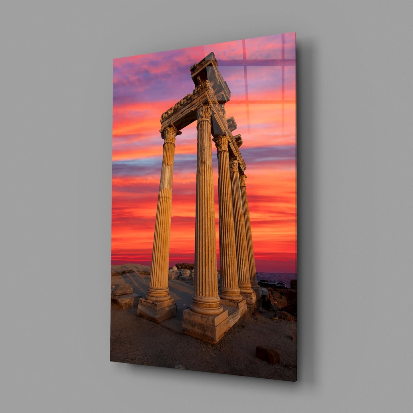 Ancient Greek columns with ornate capitals stand against a vibrant orange and pink sunset sky on glass wall art.