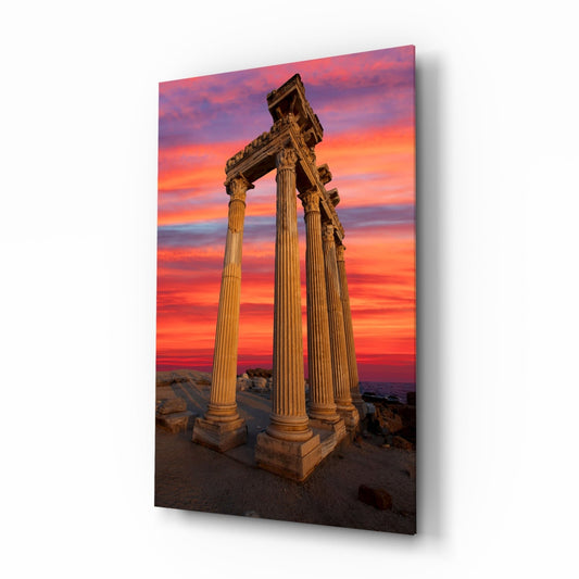Ancient Corinthian columns with ornate capitals stand against a vibrant sunset sky in shades of orange, pink, and purple.