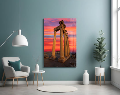 Temple of Apollo glass wall art displayed in a room setting showing ancient columns against a vibrant orange and pink sunset sky.