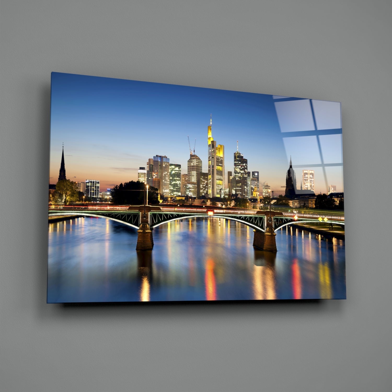 Frankfurt skyline at dusk reflected in river with illuminated buildings and iron bridge, displayed as blue-framed glass wall art.