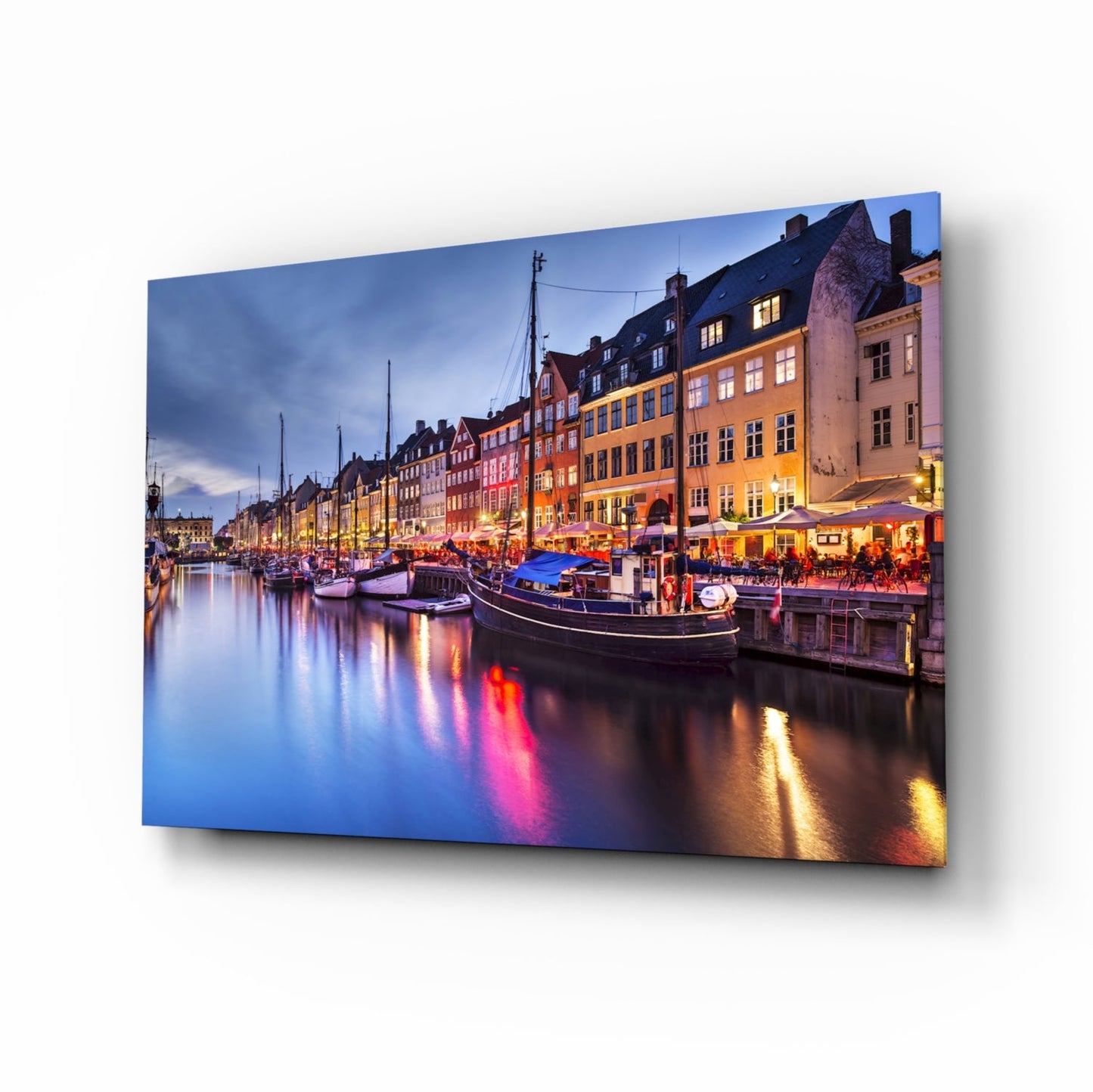 Glass wall art panel displaying twilight photo of colorful Nyhavn harbor with illuminated buildings and boats reflected in water.