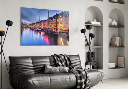 Nyhavn glass wall art displaying illuminated colorful buildings and boats reflecting in water, hung above black leather sofa in modern living room.