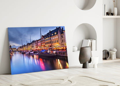 Glass wall art leaning against white wall displaying illuminated Nyhavn harbor with colorful buildings and boats at dusk.