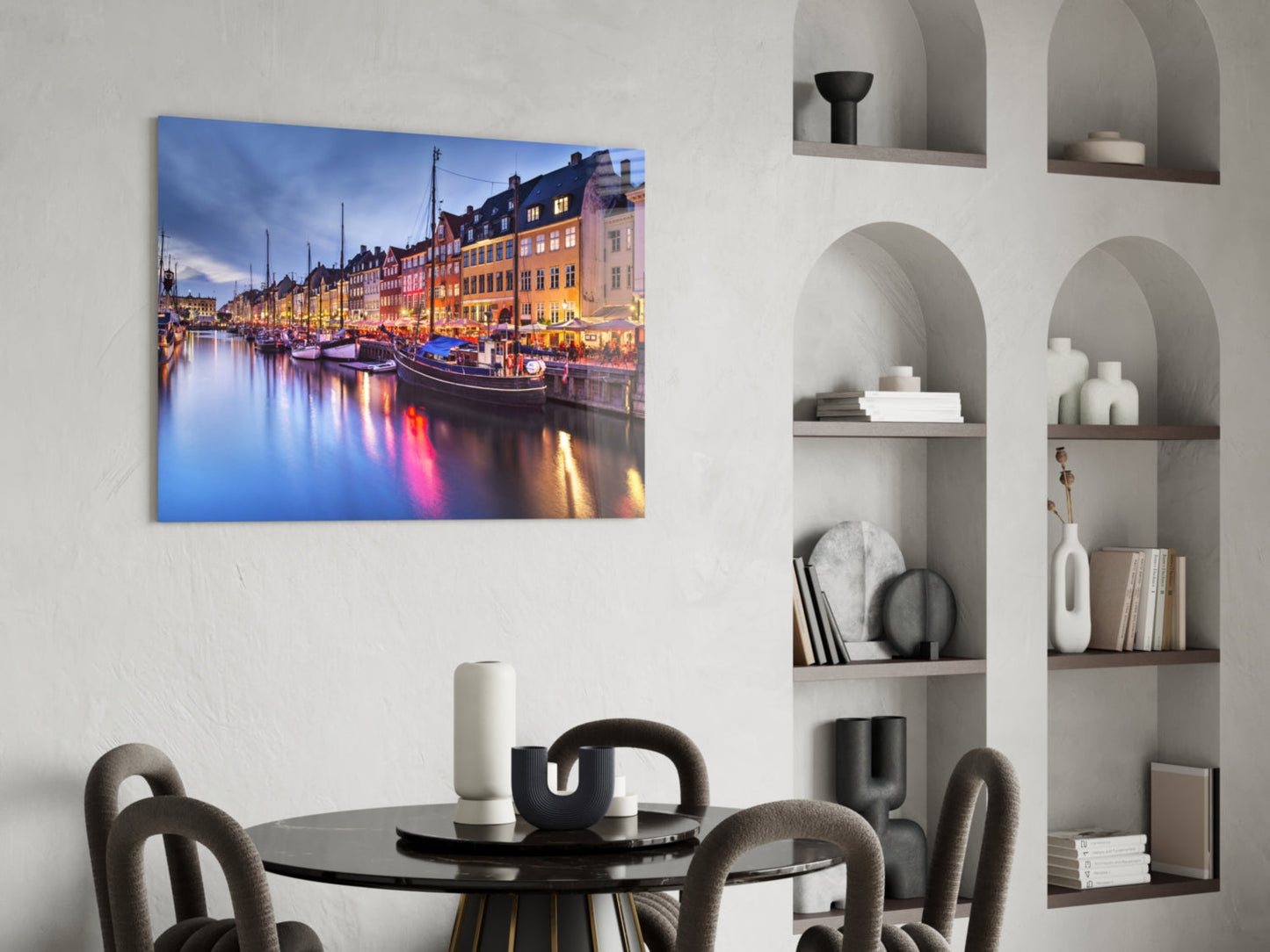 Glass wall art depicting colorful illuminated buildings and boats reflected in calm water at dusk, displayed above a round black table.
