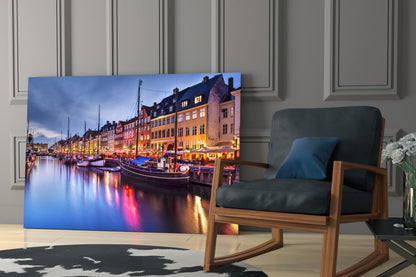 Glass wall art displaying Nyhavn harbor at twilight with illuminated colorful buildings and boats reflected in water, displayed in modern living room setting.