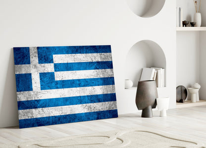 Greek flag glass wall art leaning against white wall in minimalist interior with decorative objects and shelving.