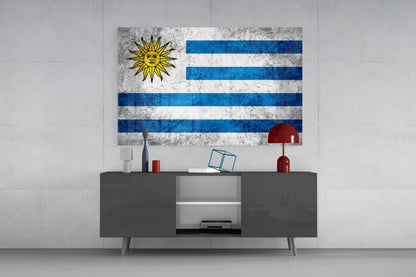 Uruguayan flag glass wall art displaying white and blue horizontal stripes with yellow sun symbol, mounted above gray media console in modern interior.