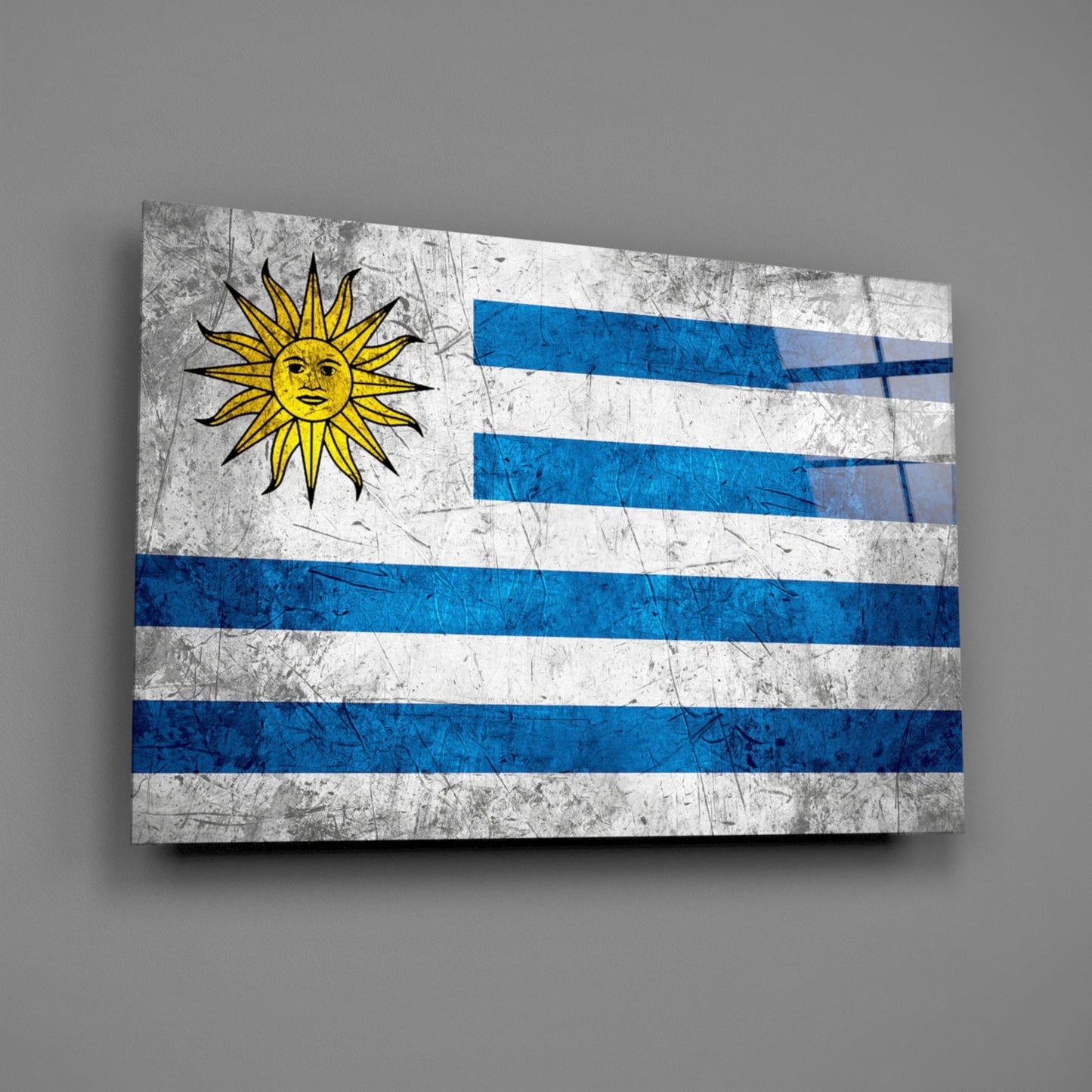 Distressed Uruguayan flag glass wall art displayed at an angle against a gray wall, showing blue and white stripes with a yellow sun symbol.