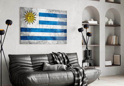 Uruguayan flag glass wall art with blue and white stripes and yellow sun displayed above black leather sofa in modern living room.