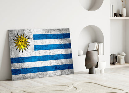 Uruguayan flag glass wall art leaning against white wall in minimalist interior with decorative objects.
