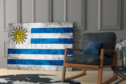 Uruguayan flag glass wall art displaying blue and white horizontal stripes and yellow sun symbol, leaning against wall beside wooden chair.