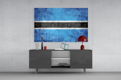 Botswana flag glass wall art displaying blue, white, and black horizontal stripes mounted above a dark gray media console with red decorative objects.