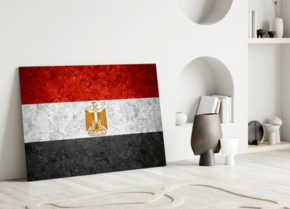 Egyptian flag glass wall art leaning against white wall in minimalist room with decorative objects.